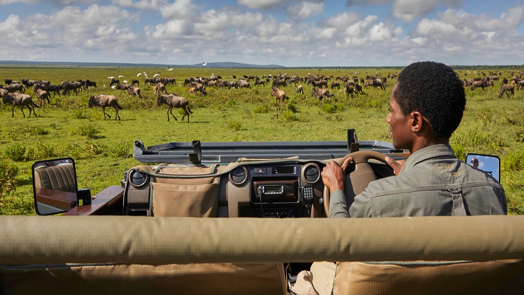 A Songa guide at the wheel of an open safari vehicle, wildebeest herds spread across the plains ahead